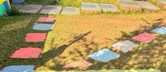 Área externa com gramado atravessado por um caminho de placas de concreto coloridas em rosa, azul, verde e bege, embutidas na grama.