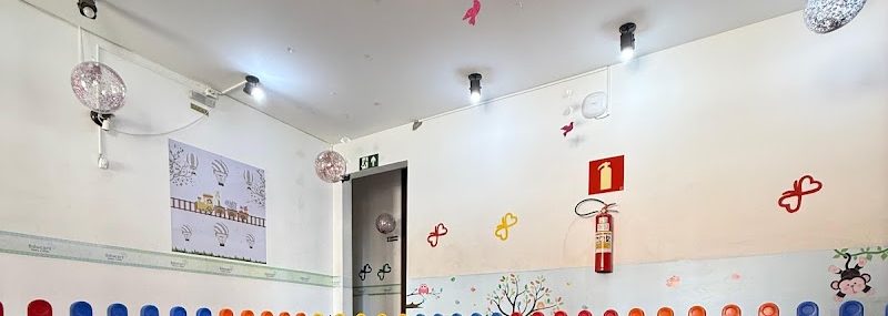 Espaço interno amplo com piso emborrachado em placas, iluminação em trilho, faixa decorativa infantil na parede, móbiles pendurados no teto e cercado de proteção colorido.
