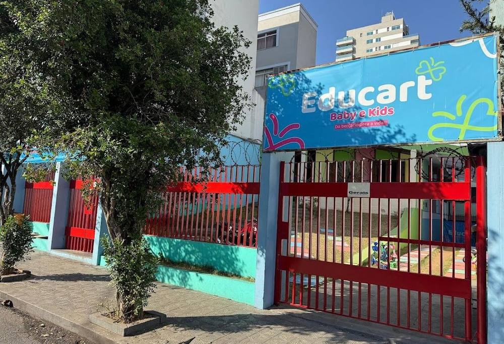 Fachada da Educart Baby e Kids: muro em turquesa e vermelho, portão de grade vermelha e banner azul com o nome da escola e a frase Do berçário para a vida.