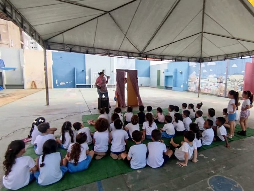 Pátio coberto de grande vão, com cobertura de lona sobre estrutura metálica, piso com circuito pintado, traves de gol e murais coloridos nas paredes. No centro, tapetes de grama sintética onde as turmas se reúnem.