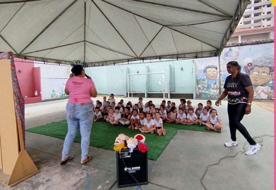 Outro ângulo do pátio coberto, mostrando a estrutura da cobertura, murais artísticos de grande formato com rostos infantis e um teatro de fantoches montado para as crianças.