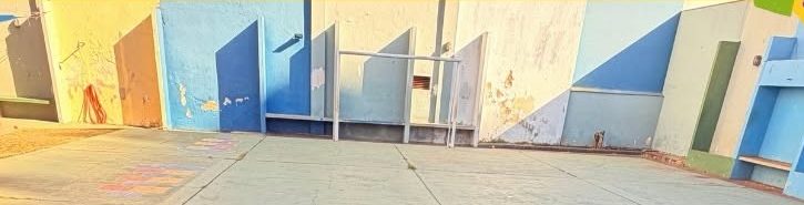 Pátio descoberto com piso de concreto e demarcações pintadas, parede alta pintada em tons de azul e branco, bancos de alvenaria embutidos e um escorregador ao fundo.