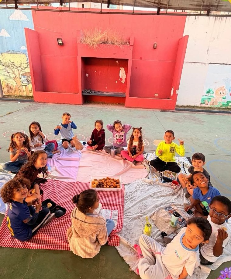 Crianças sentadas em toalhas de piquenique no pátio da escola, com lanches compartilhados, murais pintados ao fundo e brinquedo de escalada ao lado.
