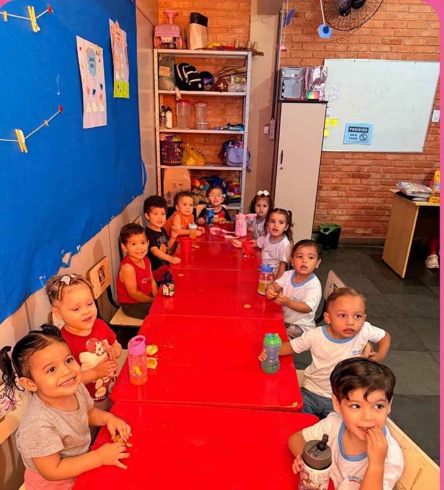 Sala do maternal na hora do lanche: mesas infantis vermelhas enfileiradas, parede de tijolo aparente, painel azul com trabalhos das crianças pendurados em um varal e lousa branca com a mensagem sejam bem-vindos.