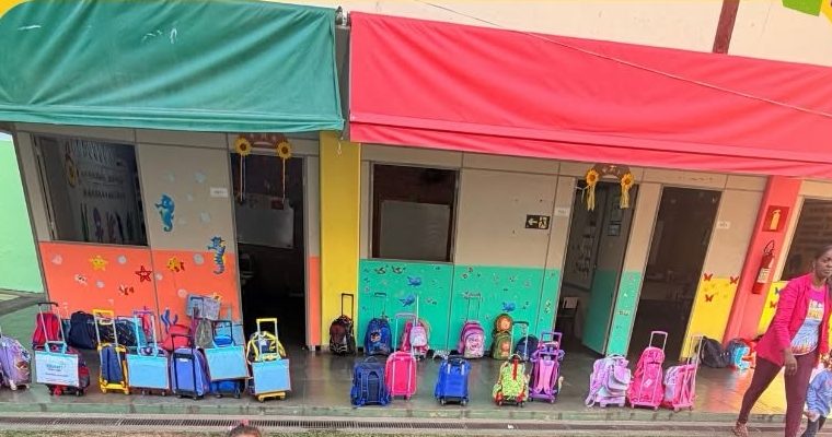 Varanda coberta de acesso às salas, com toldos verde e vermelho, portas de alumínio, painéis decorativos temáticos nas paredes e uma fileira de mochilas de rodinha organizadas no corredor.